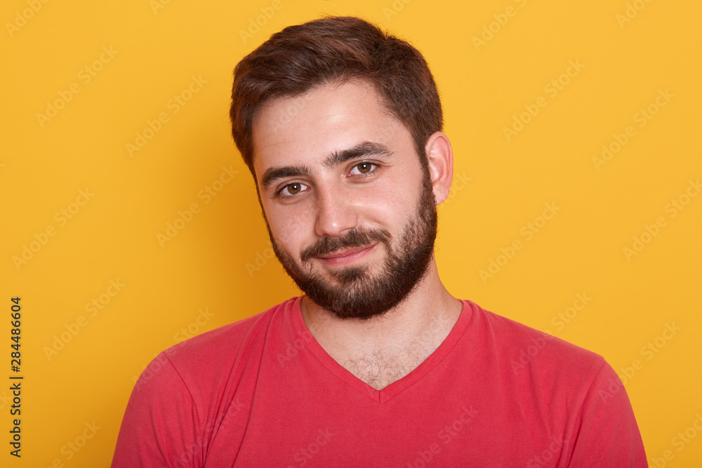 Close up portrait of young handsome man with beard wearing red casual t shirt posing isolated over yellow background, attractive male has calm facial expression, looks smiling directly at camera. Close up portrait of young handsome man with beard wearing red casual t shirt posing isolated over yellow background, attractive male has calm facial expression, looks smiling directly at camera.