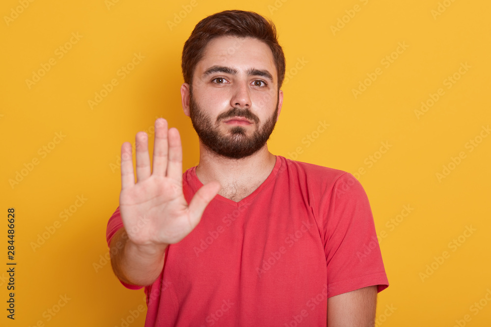 Portrait of calm bearded young man wearing red casual t shirt standing with stop warning gesture hand and looking at camera with serious face, handsome male posing isolated over yellow background. Portrait of calm bearded young man wearing red casual t shirt standing with stop warning gesture hand and looking at camera with serious face, handsome male posing isolated over yellow background.