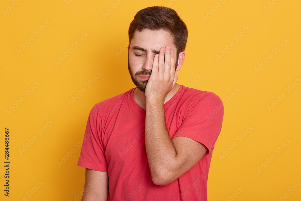 Exhausted young unshaven ing half of face with hand while standing against yellow studio background, looks tired, feeling depressed, has problems, posing with closed eyes, dressed red t shirt. Exhausted young unshaven ing half of face with hand while standing against yellow studio background, looks tired, feeling depressed, has problems, posing with closed eyes, dressed red t shirt.