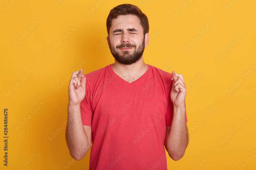 Close up portrait of handsome young man wearing red t shirt keeping eyes closed and fingers crossed while standing against yellow background, waiting results of exam, wants his dream comes true. Close up portrait of handsome young man wearing red t shirt keeping eyes closed and fingers crossed while standing against yellow background, waiting results of exam, wants his dream comes true.