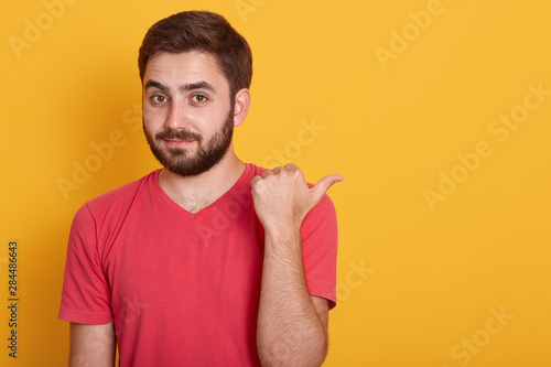 Close up portrait of handsome young male with beard pointing aside with thumb and looking at camera, unshaven man posing dressed in red casual t shirt. Copy space for promotin text or advertisment.