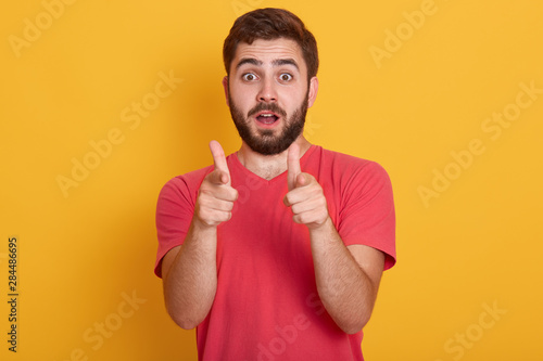 Image of handsome young man standing isolated over yellow background, looking at camera and pointing with fore fingers, wearing red casual t shirt, posing with open mouth. People emotions concept.