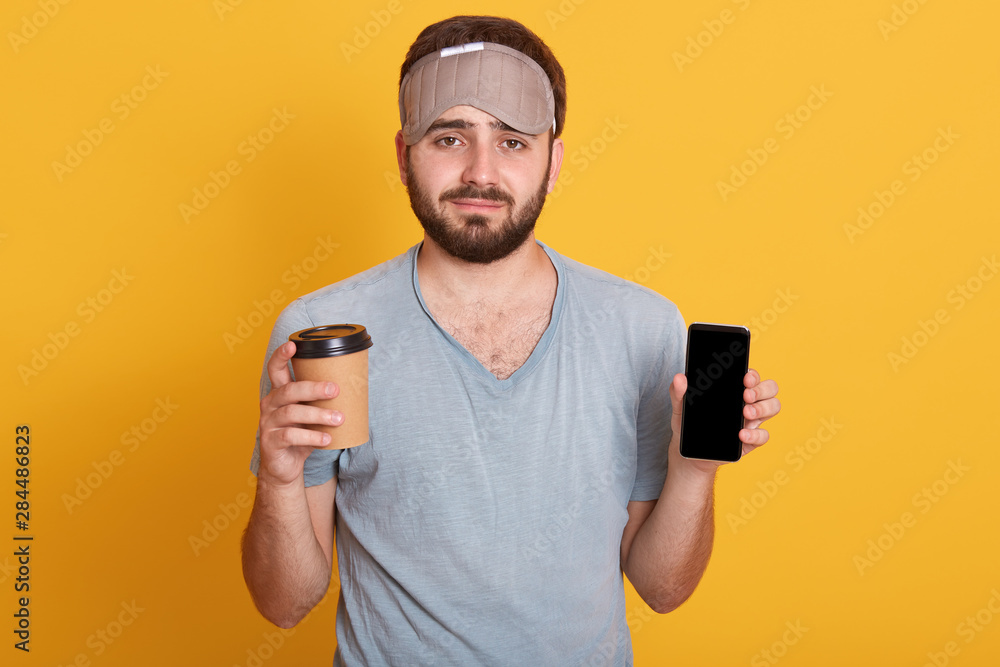Portrait of upset handsome bearded young hipster man in gray t shirt and blindfold standing with coffee in one hand and phone with blank screen in another, isolated over pink studio background. Portrait of upset handsome bearded young hipster man in gray t shirt and blindfold standing with coffee in one hand and phone with blank screen in another, isolated over pink studio background.
