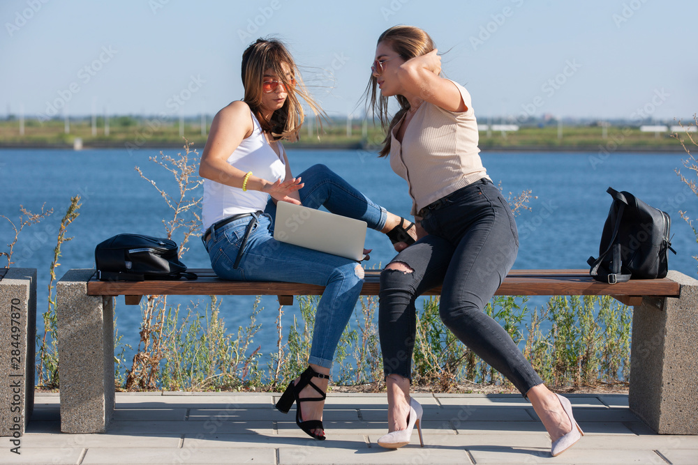 Two young female girlfriends having fun while sitting on a bench while one showing to screen of a laptop while another one is laughing outside.