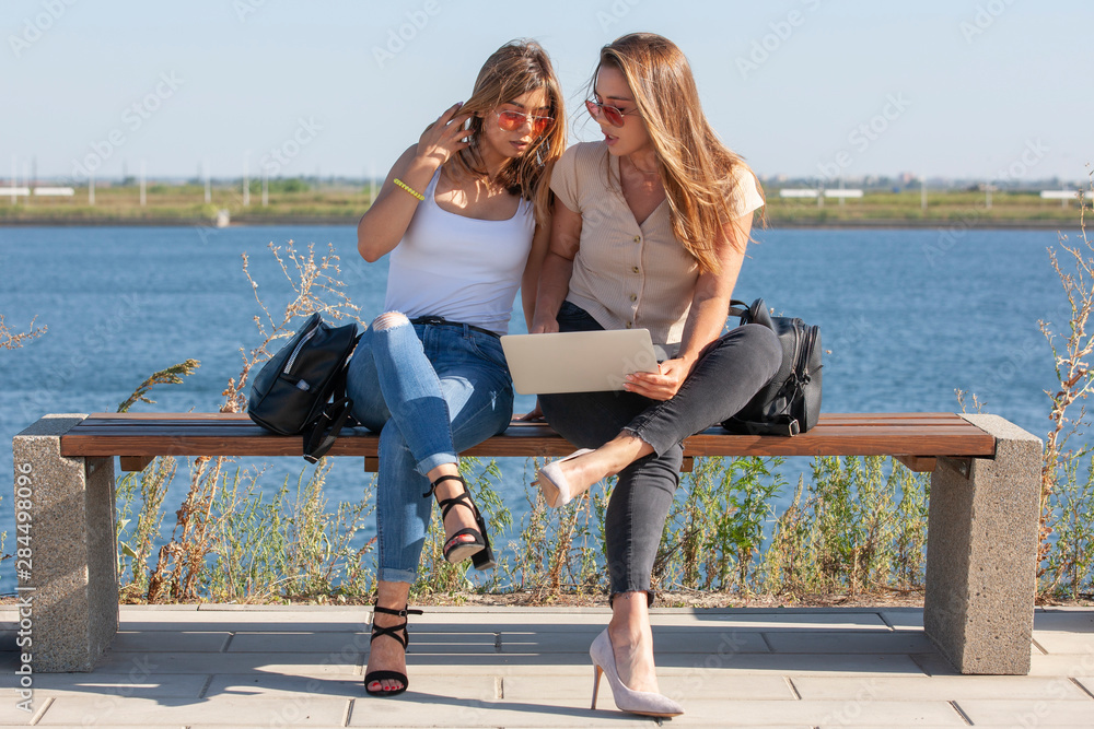 Two girls sit on a bench in the summer park. They look at the laptop screen.