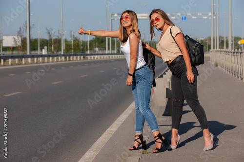 Two beautiful young women are hitchhiking on the road and try to find someone to take them to their hotel.