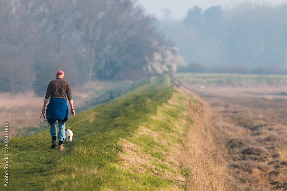 A middle aged man walking his dog off the lead along a rural footpath next to the river Deben in Suffolk
