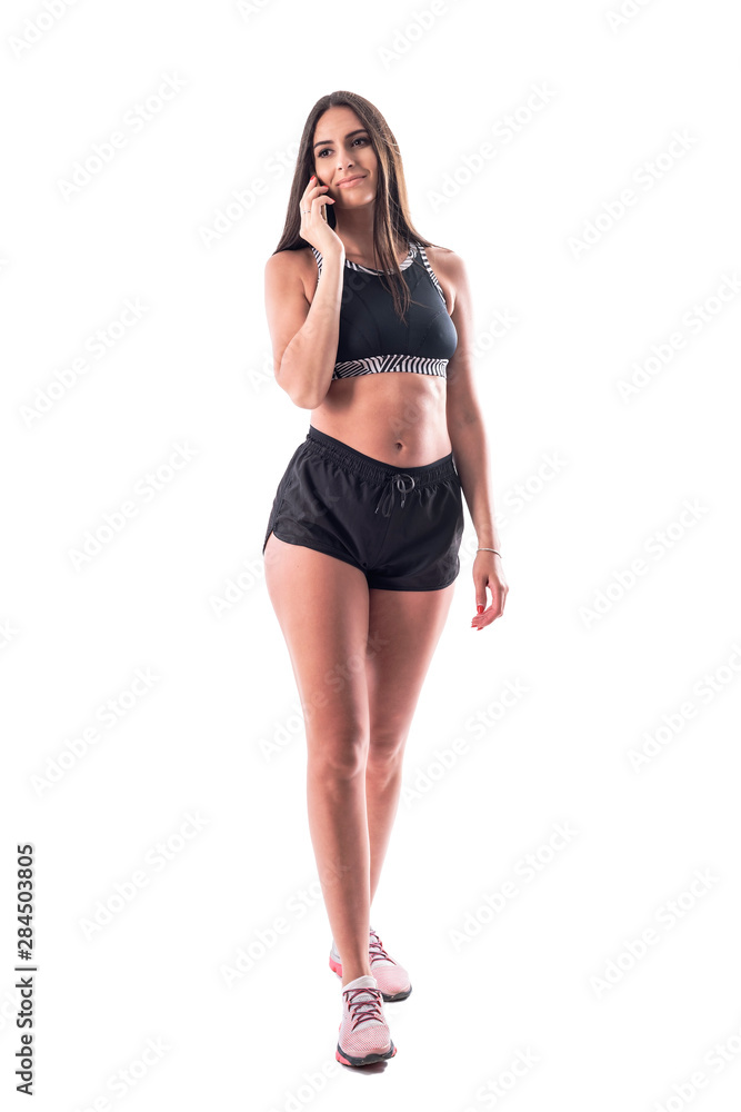 Fit sporty attractive gym woman walking and talking on the cellphone looking away. Full body isolated on white background. Fit sporty attractive gym woman walking and talking on the cellphone looking away. Full body isolated on white background.