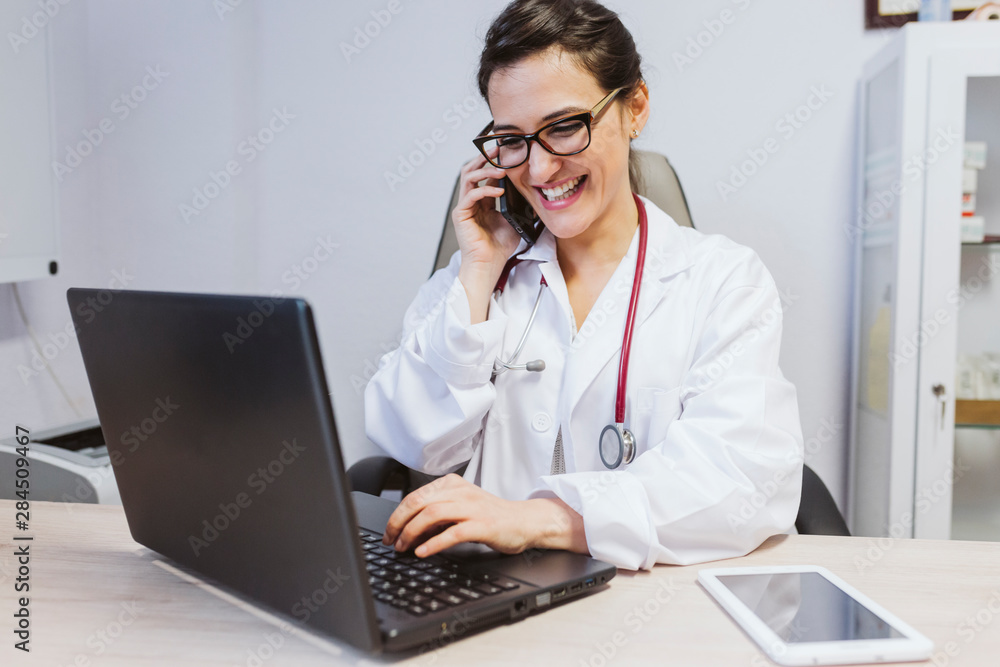 young doctor woman working on laptop at the consult. Talking on mobile phone. Modern Medical concept indoors young doctor woman working on laptop at the consult. Talking on mobile phone. Modern Medical concept indoors