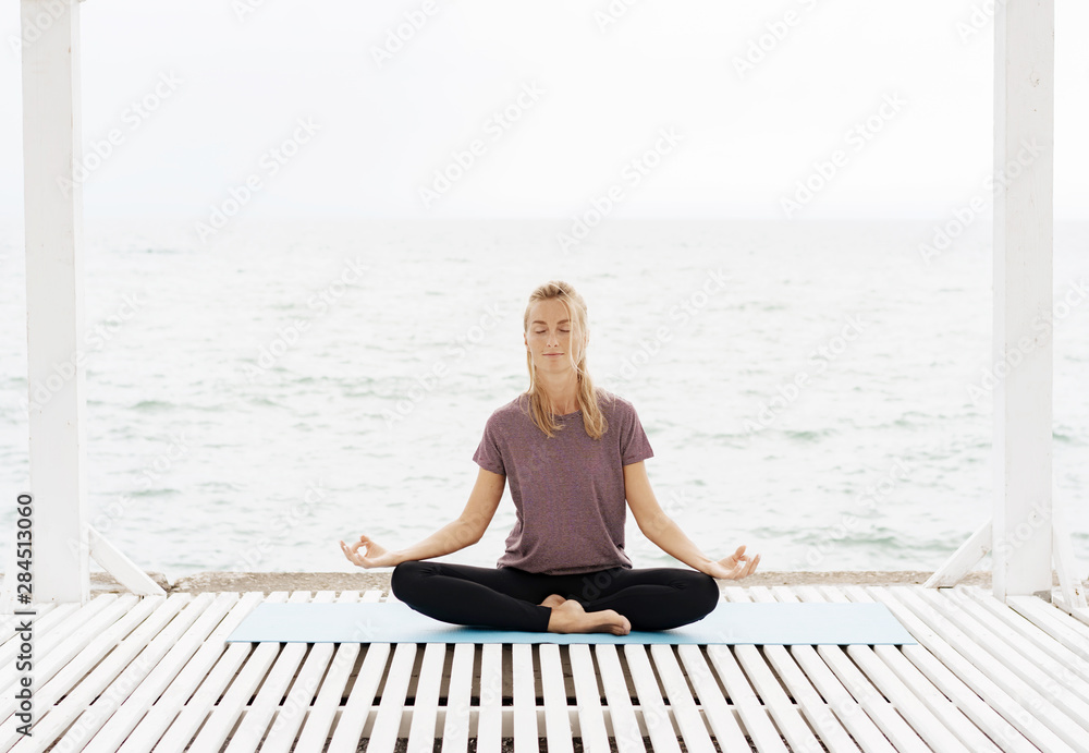 Caucasian blonde woman meditates sitting in lotus position on the seashore, concept of physical and mental health. Place for an inscription. Motivational image.