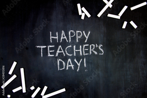 Back to school, college, university concept. Congratulatory inscription of happy teachers day on blackboard background from pupils. Pieces of white chalk are scattered on black canvas.