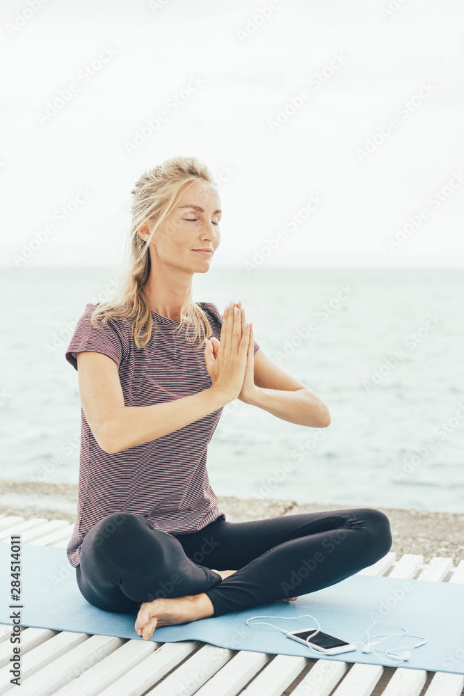 A Caucasian blonde woman in lotus position greets the namaste before starting yoga practice. Sports concept. Vertical image. Physical and mental health.
