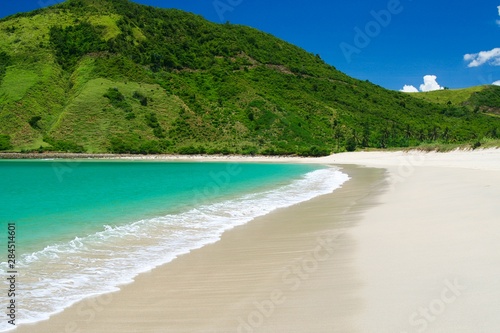 Isolated lonesome lagoon with turquoise sea  white surf in shallow water  white soft sand and lush green hill on Lombok  Indonesia