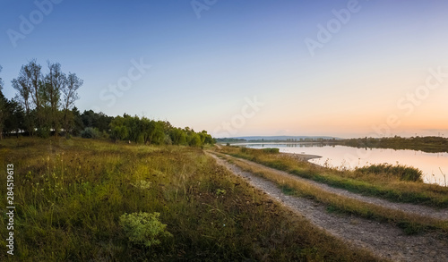 Rural panoramic landscape as a country road separates the lake from the forest. Beautiful evening scene, calm autumn background near a meadow of steppe vegetation.