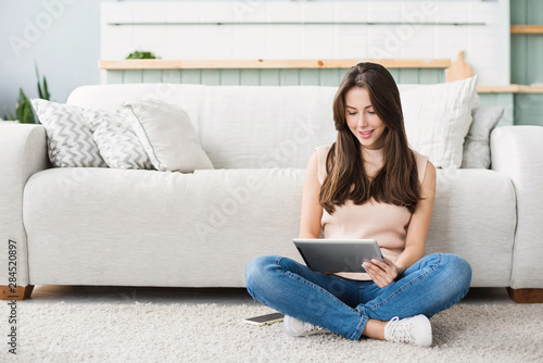 Beautiful young woman with digital tablet relaxing at home. Happy smiling girl sitting in living room using tablet pc. Resting, relaxation, technology concept