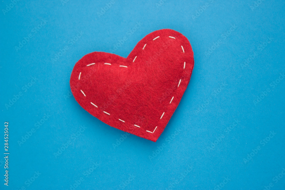 Fototapeta premium Red heart fabric with white threads on a blue background Love, romance concept. Felt bright hearts with embroidered lines.