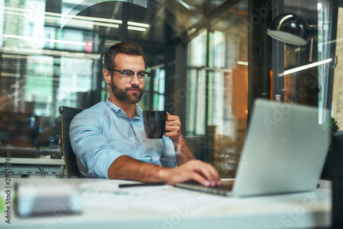 Morning coffee. Portrait of young and successful bearded man in eyeglasses holding cup of coffee and working with laptop while sitting at his working place