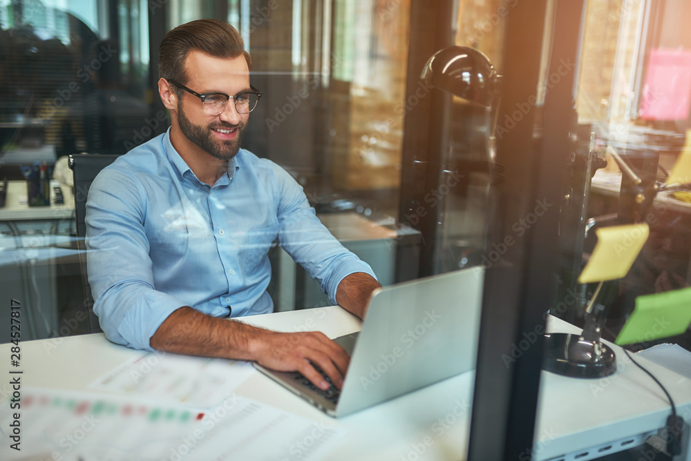 Fototapeta premium Great results Young bearded businessman in eyeglasses and formal wear looking at laptop and smiling while sitting in the modern office