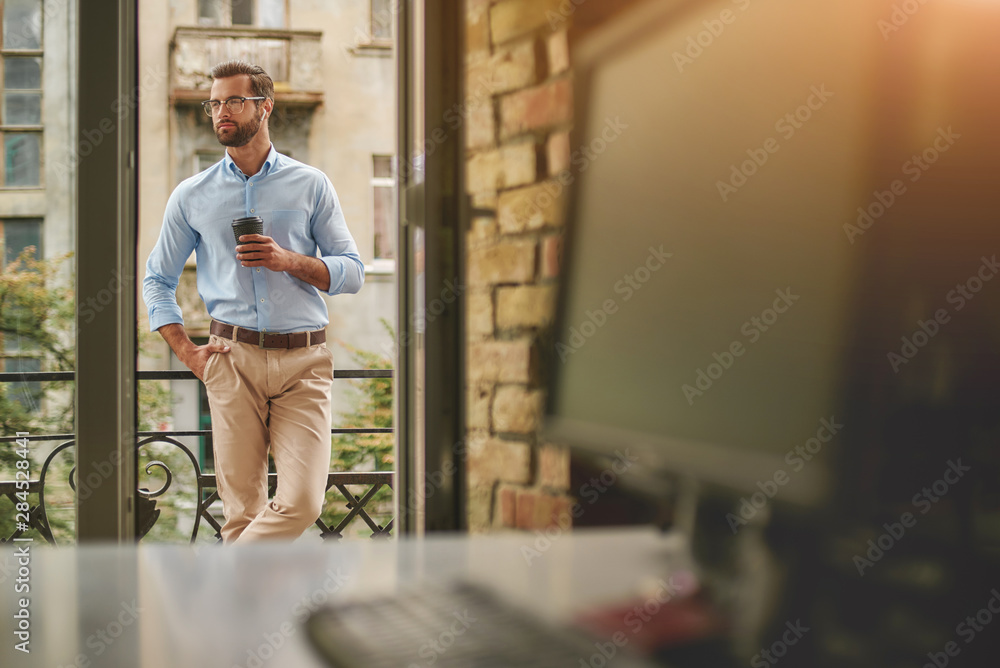 Having coffee break. Young and handsome bearded man in eyeglasses and formal wear holding cup of coffee and looking away while standing at the office balcony - obrazy, fototapety, plakaty 