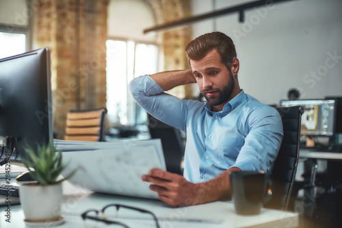 I am not sure. Frustrated bearded engineer in formal wear looking at blueprint while sitting in the modern office