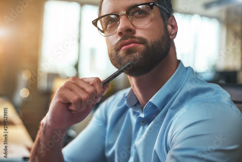 Lost in thougths. Portrait of young bearded man in eyeglasses and formal wear looking away and thinking while working in the modern office photo
