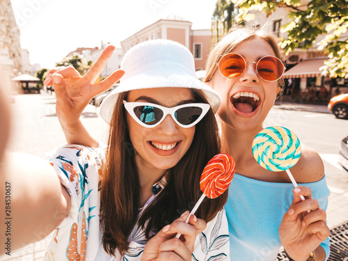 Two young smiling hipster women in casual summer clothes. Girls taking selfie self portrait photos on smartphone.Models posing on street background in sunglasses and panama hat.Eating candy lollipop
