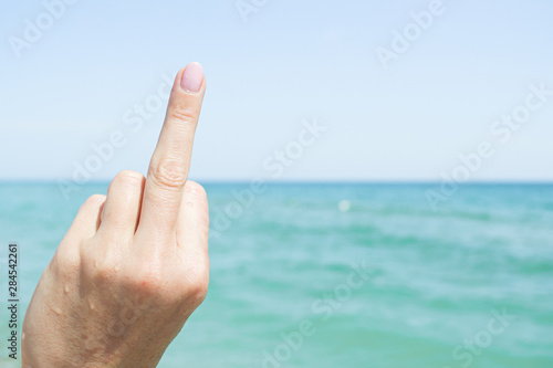 Sign with the middle finger of a female hand on a background of blue sky and blue sea