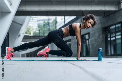Young female in sportswear doing core workout on fitness mat at city street in urban zone