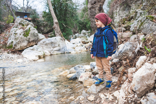 A child with a backpack stands near a mountain river.