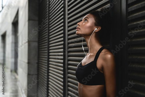 Side view of sporty woman listening to music on earphones and leaning to a wall outdoors taking break after workout