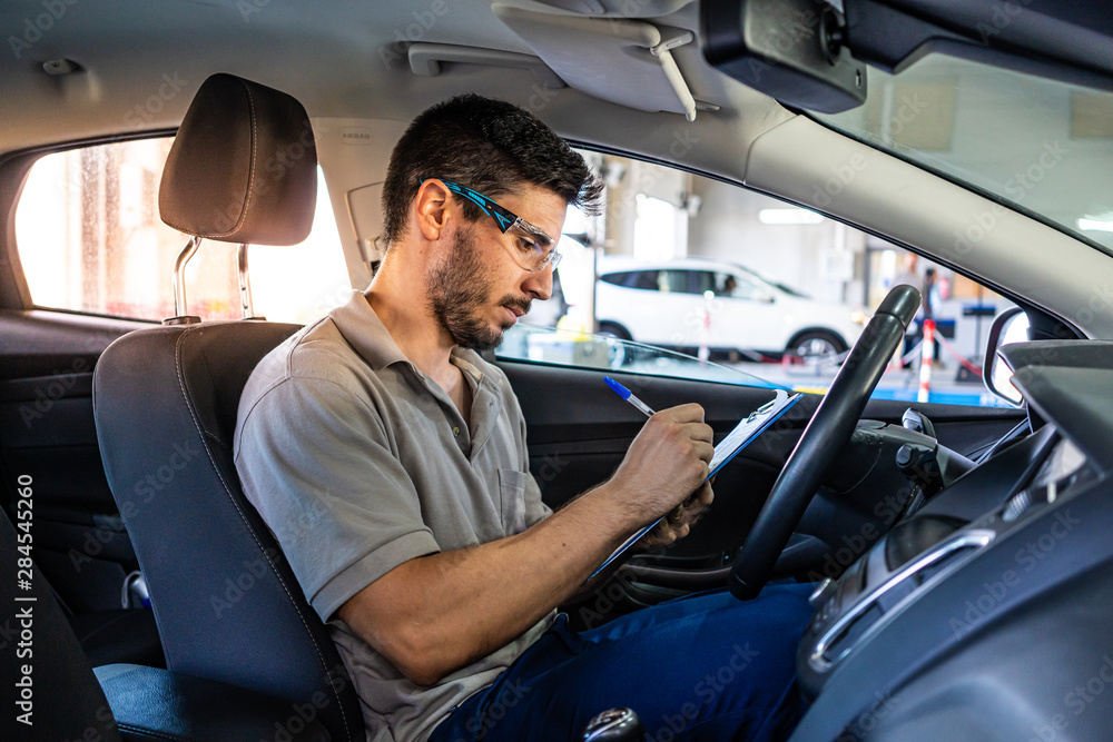 Technician with safety glasses sitting in a car seat checking a list during a vehicle inspection