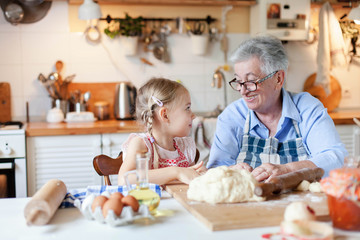 Senior woman and child make dough for bread together. Family is cooking in cozy kitchen at home. Grandmother is teaching little girl. Cute kid is helping to prepare meal for Thanksgiving Day dinner.