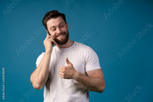 casual man showing thumb up sign while talking on the telephone and smiling for the camera. isolated on blue background