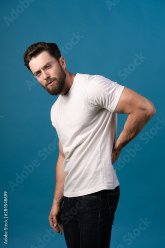Young male suffering from back pain against a blue background. Portrait of a handsome man. Vertical photo. Concept- backpain, spine problems.
