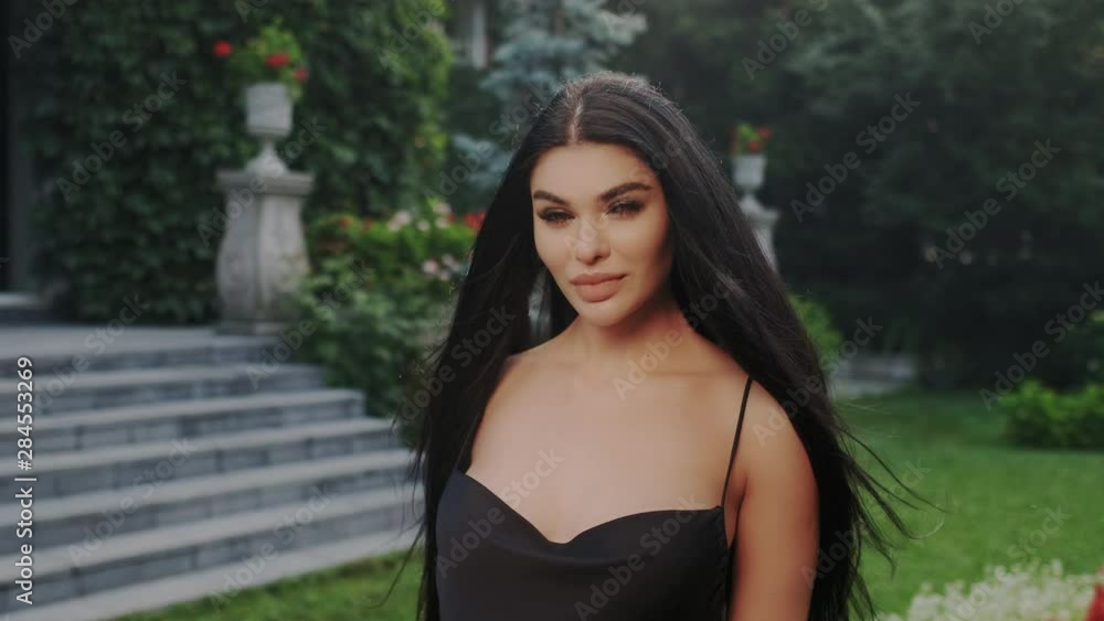 An attractive sexy luxurious brunette stands in the garden in a beautiful black evening dress with an open neckline. Hair sways in the wind. Girl's face after cosmetological procedures. An attractive sexy luxurious brunette stands in the garden in a beautiful black evening dress with an open neckline. Hair sways in the wind. Girl's face after cosmetological procedures.