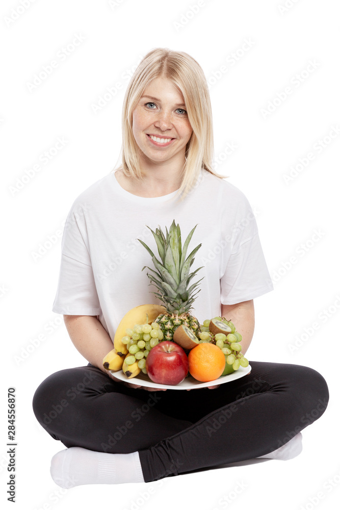 A smiling young woman is sitting with a plate of fruit in her hands. Isolated over white background. Vertical. A smiling young woman is sitting with a plate of fruit in her hands. Isolated over white background. Vertical.