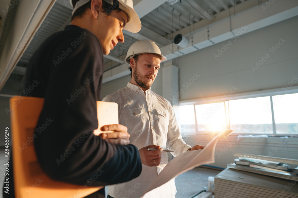 Building industry. Young engineer discussing with building worker on construction site. Industrial engineers have meeting while uses blueprint Building industry. Young engineer discussing with building worker on construction site. Industrial engineers have meeting while uses blueprint