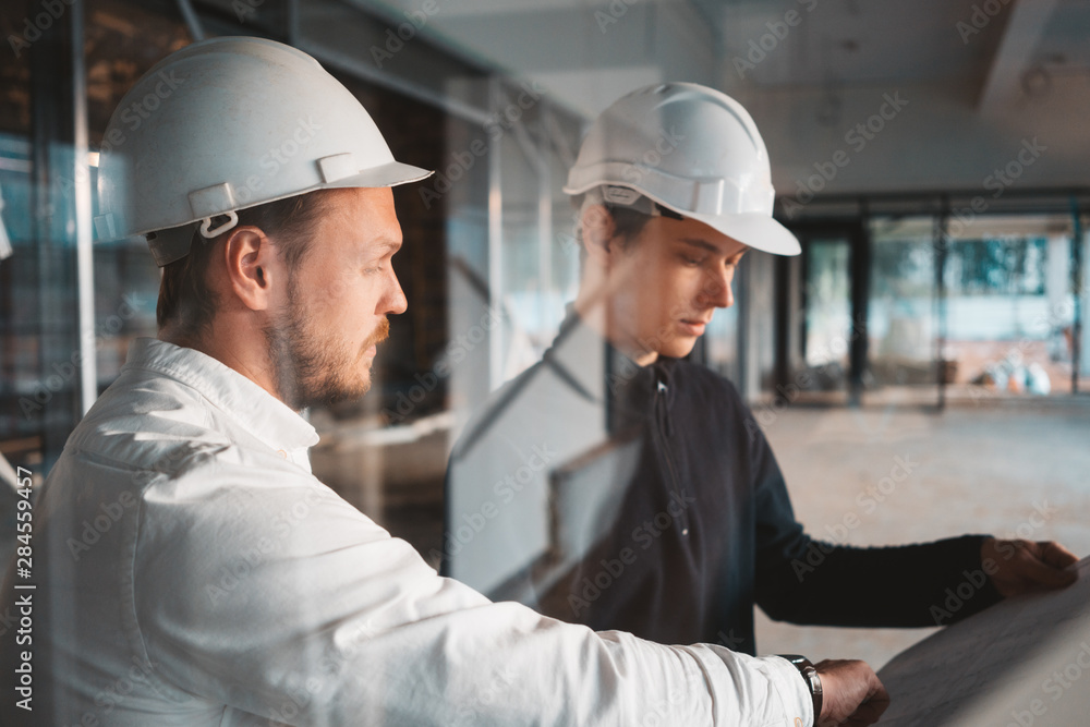 Building worker and architect discussing build drawing on construction site. Two industrial engineers wearing safety hard hat have meeting on commercial building structure