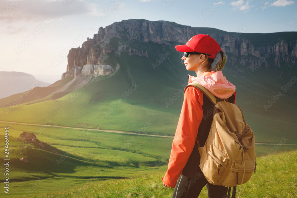 Girl athlete in a red cap sunglasses and a yellow backpack stands on a green slope against the background of the epic cliffs of the Caucasus
