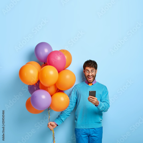 Overjoyed surprised man focused in smartphone device, pleasantly shocked to get message from former girlfriend on birthday, sends invitation to friends on party, carries bunch of colorful balloons