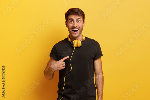 Cheerful delighted man points at himeself, asks whether someone choosed him, has dark hair, dressed in black t shirt, models over yellow background, wears modern headphones on neck for listening music