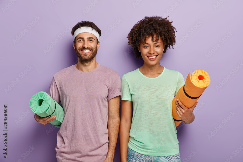 Indoor shot of happy mixed race female and male ready for fitness training, carry rolled up mats under arms, have glad faces, enjoy active life and everyday regular workout, wear casual sport clothes - obrazy, fototapety, plakaty 