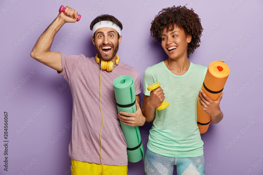 Photo of happy guy and woman work on biceps with weights, carry karemats, have joyful expressions, enjoy training together, dressed in casual wear, being motivated for healthy lifestyle and sport Photo of happy guy and woman work on biceps with weights, carry karemats, have joyful expressions, enjoy training together, dressed in casual wear, being motivated for healthy lifestyle and sport