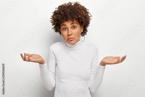 People, attitude and life perception concept. Clueless unaware woman with curly hair, spreads palms in doubt, expresses uncertainty, has hesitant expression, wears white jumper, shrugs shoulders