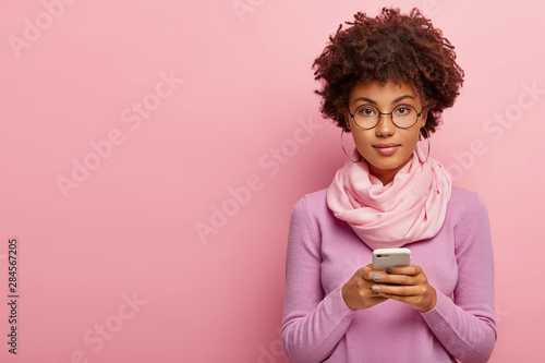Beautiful young woman with curly dark hair, stays in touch, uses modern gadget, makes her own blog, connected to wireless internet, wears glasses, big hoop earrings, scarf and turtleneck poses on pink