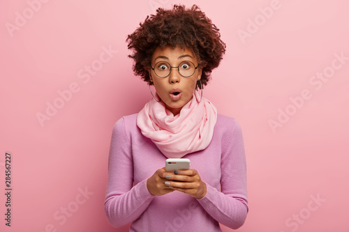 Emotive surprised dark skinned Afro female blogger makes publicaion on mobile phone, shocked to get unexpected message, keeps mouth opened, looks through optical glasses, wears stylish outfit