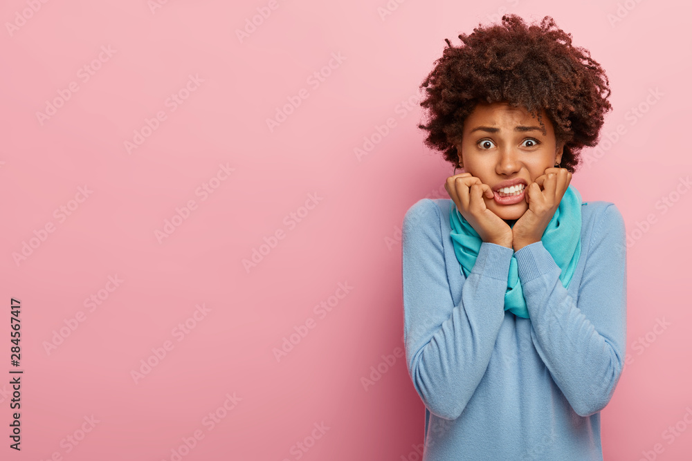 Nervous displeased woman bites finger nails, has upset expression, being under pressure and panic, wears casual blue outfit, has crisp hair, stands against pink studio wall free space aside for advert Nervous displeased woman bites finger nails, has upset expression, being under pressure and panic, wears casual blue outfit, has crisp hair, stands against pink studio wall free space aside for advert