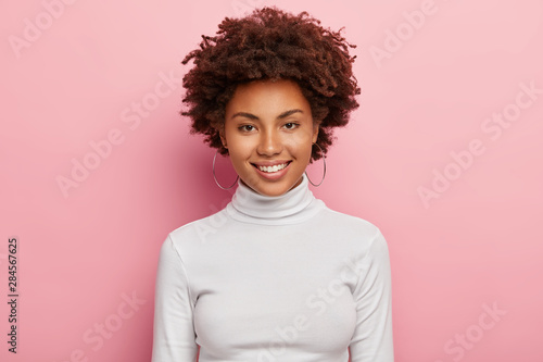 Gorgeous lovely woman has pleasant smile on face, healthy skin, Afro haircut, wears casual white turtleneck jumper, round earrings, looks happily directly at camera, has no make up, feels joyful