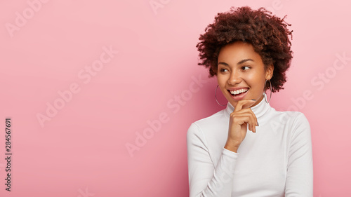 Shot of beautiful smiling woman touches chin gently, smiles broadly, being in perfect mood, notices pleasant scene aside, wears casual white turtleneck, isolated over rosy background, free space