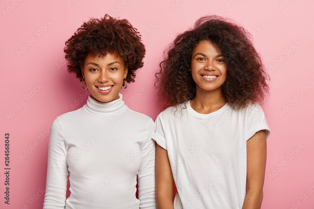 Portrait of beautiful two female models happy to meet, make photo against pink background, smile broadly, express good emotions, look similar, have Afro hair, wear white turtleneck and t shirt Portrait of beautiful two female models happy to meet, make photo against pink background, smile broadly, express good emotions, look similar, have Afro hair, wear white turtleneck and t shirt
